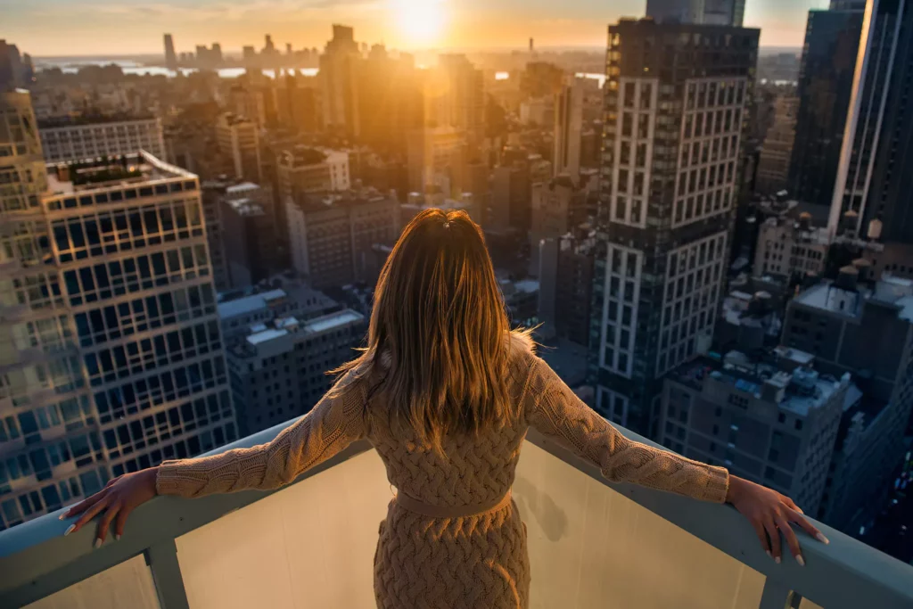 Woman with brown hair wearing light brown sweater dress looking at the Manhattan luxury real estate market skyline at sunset